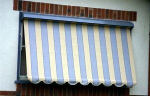 Blue and cream striped automatic roll-up canvas window awning with hood and side guide arms on a brick home